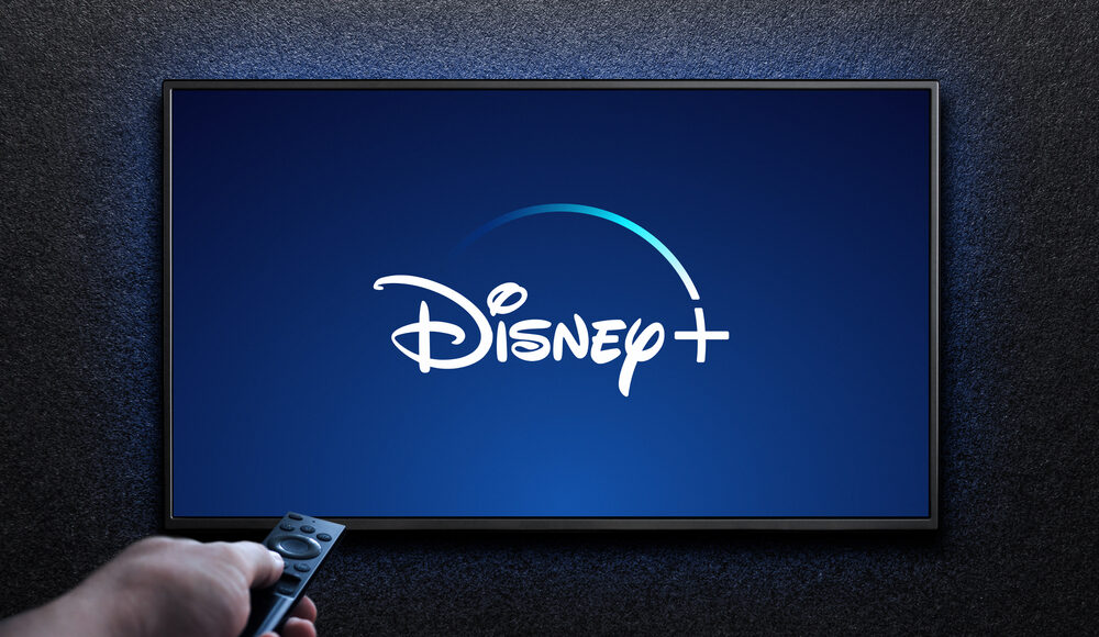 Una mano che tiene in mano un telecomando indica un televisore su cui è ben visibile il logo Disney+, illuminato su uno sfondo scuro.