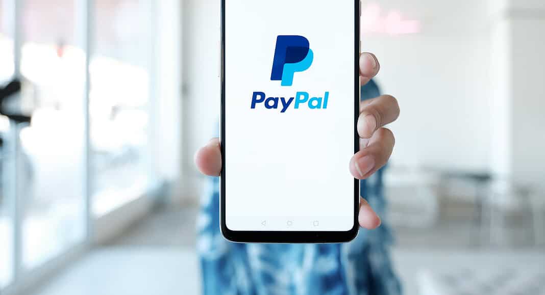 Una persona tiene in mano uno smartphone con il logo PayPal ben visibile sullo schermo in una stanza ben illuminata.