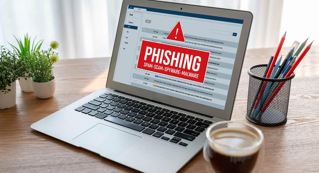 Un laptop che mostra un avviso di phishing è appoggiato su un tavolo, circondato da piante, matite e una tazza di caffè. L'avviso mette in guardia dalle e-mail sospette, assicurando che la tua casella di posta Gmail rimanga sicura e protetta.