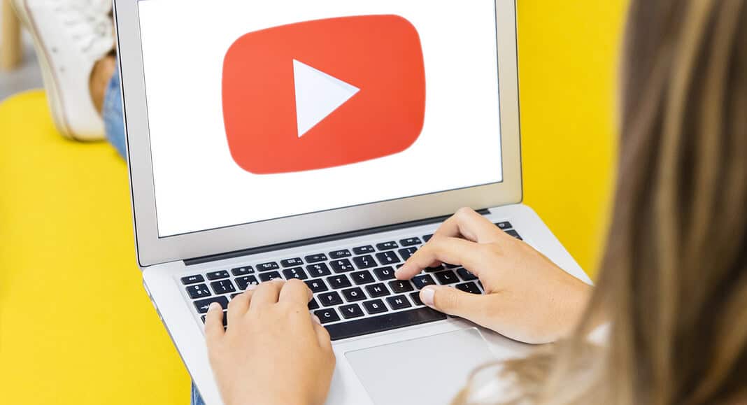 Una persona è seduta tranquillamente su un divano giallo, intenta a usare il suo computer portatile su cui è esposto l'iconico logo di YouTube.