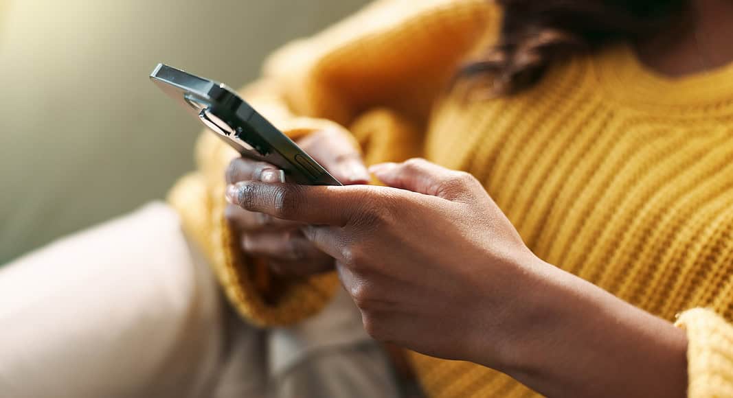 Una persona con un maglione giallo è seduta su un divano e tiene uno smartphone con entrambe le mani, probabilmente mentre controlla WhatsApp.