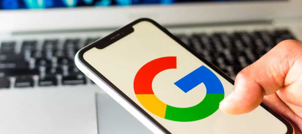 Una persona tiene in mano uno smartphone con il logo di Google, mentre Jarvis elabora i dati su un computer portatile lì vicino, su cui vengono visualizzati vari grafici finanziari.