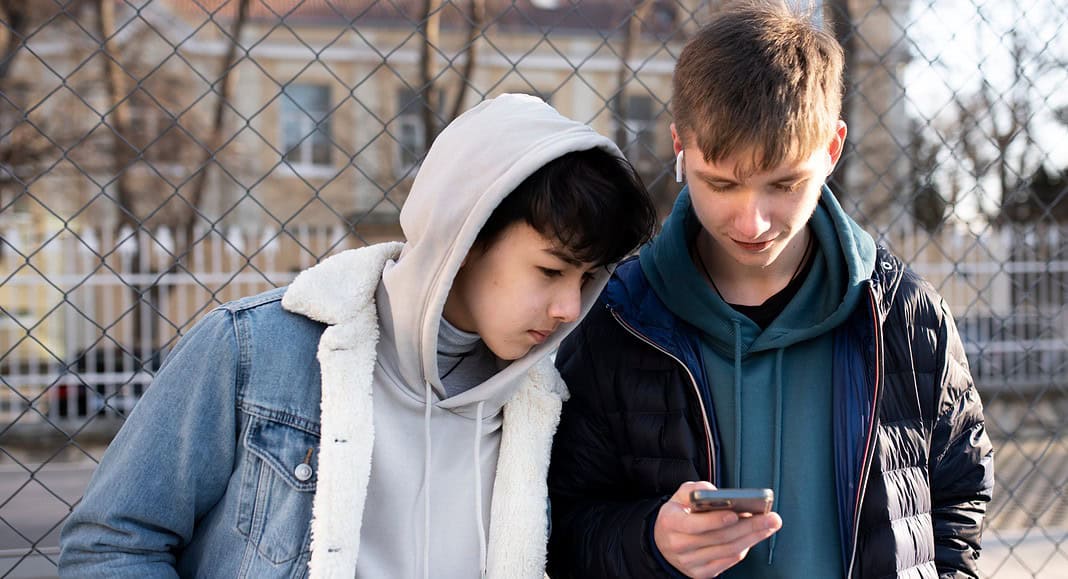 Due giovani uomini sono in piedi all'aperto, uno dei quali è impegnato socialmente mentre mostra all'altro qualcosa di interessante sul suo telefono, di fronte a una recinzione di filo spinato.