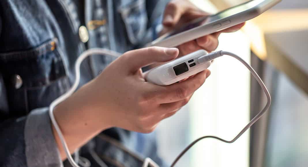 Una persona indossa una giacca di jeans e tiene in mano uno smartphone Apple e un power bank collegati tramite un cavo.